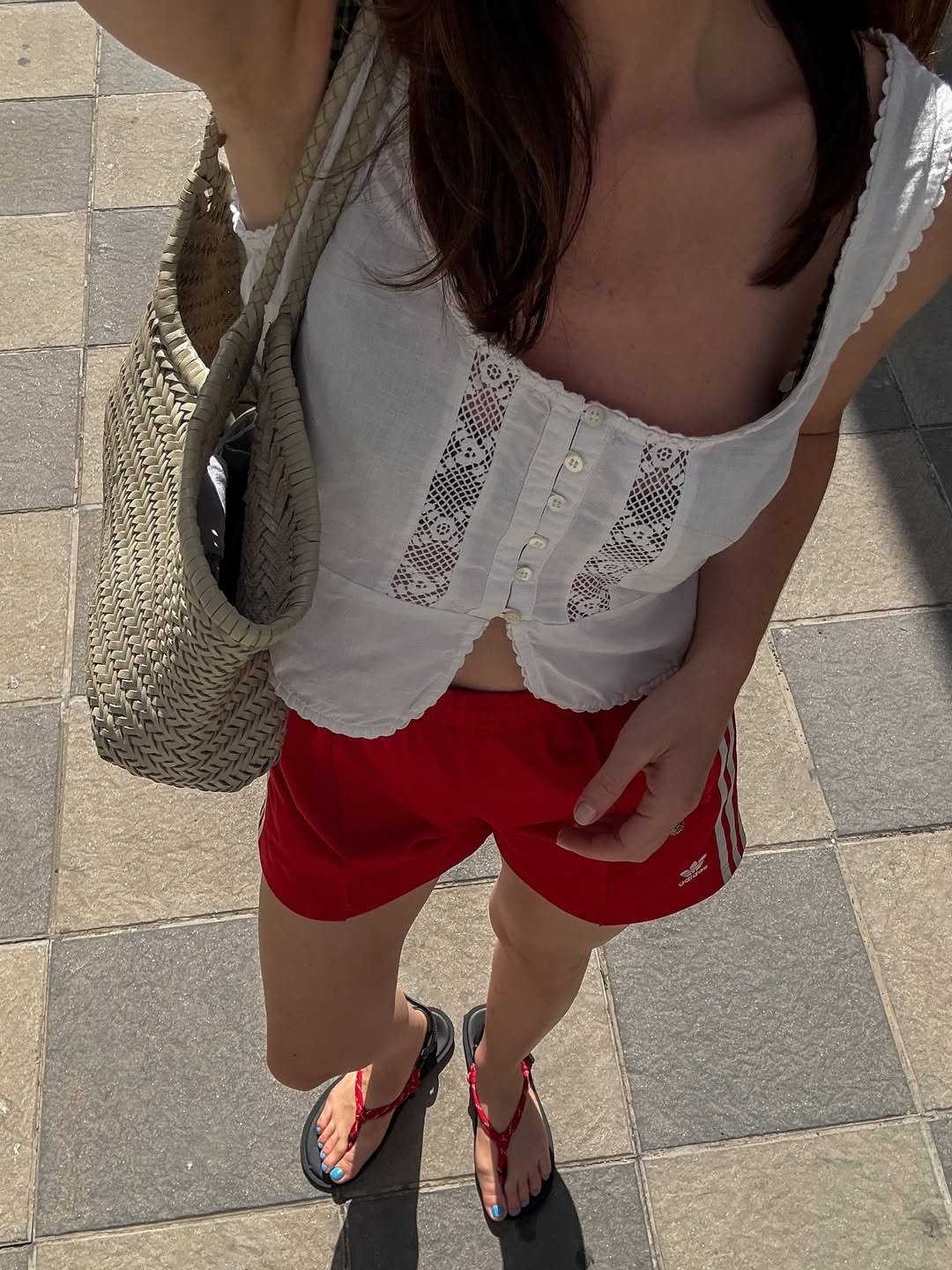 Modelo posa ao ar livre com short soltinho vermelho esportivo, regata branca rendada e bolsa de palha, em look casual e
