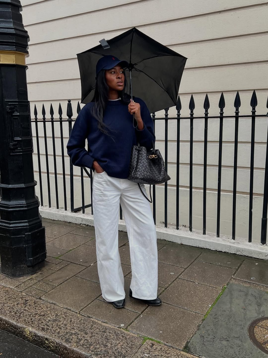 Modelo posa com tricô leve azul marinho de malha fina, calça branca ampla, bolsa preta texturizada e guarda-chuva em dia