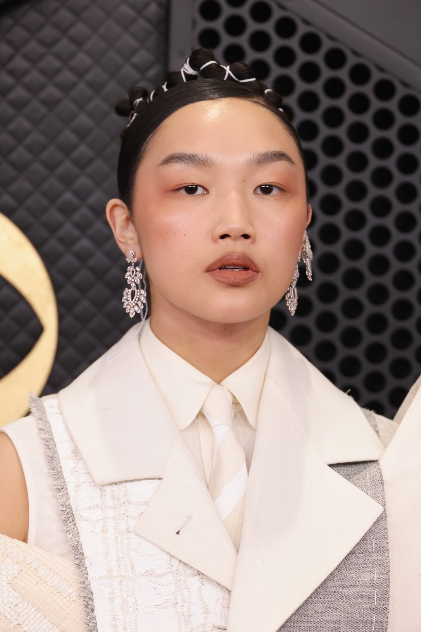 Modelo posa com expressão serena usando colete estruturado em tons off-white e cinza, gravata listrada e brincos de strass,