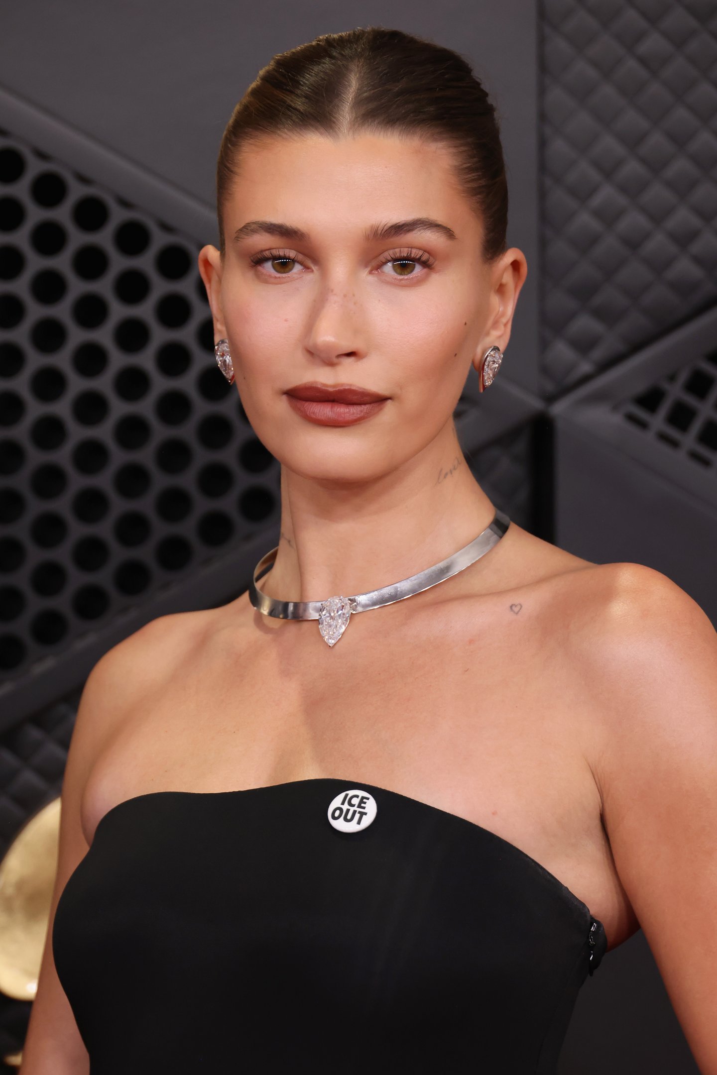 Look para Grammy 2026 com vestido tomara que caia preto, colar metálico com pedra, brincos de cristal e broche "ICE OUT",