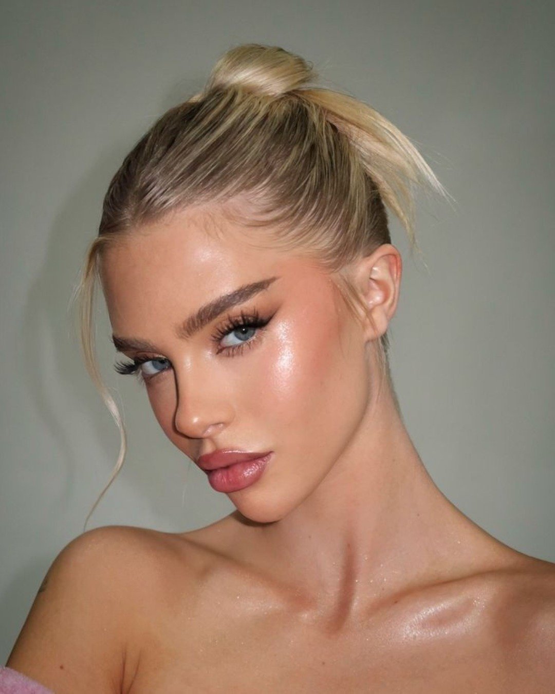 Modelo loira com coque alto posa evidenciando iluminador com efeito molhado em pele natural e olhos azuis marcantes.