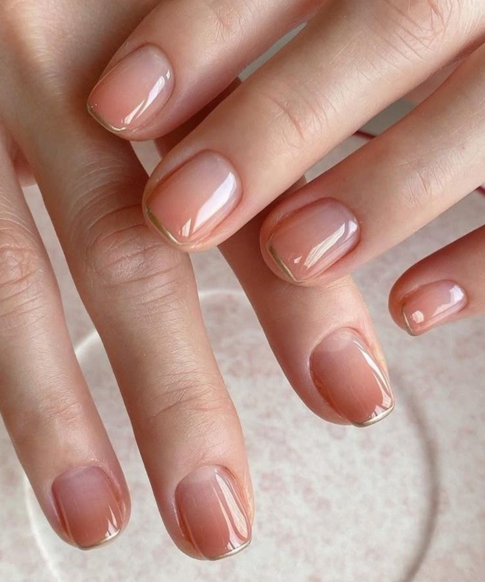 Unhas minimalistas com esmalte nude glossy e contorno dourado delicado valorizam a elegância sutil e moderna.