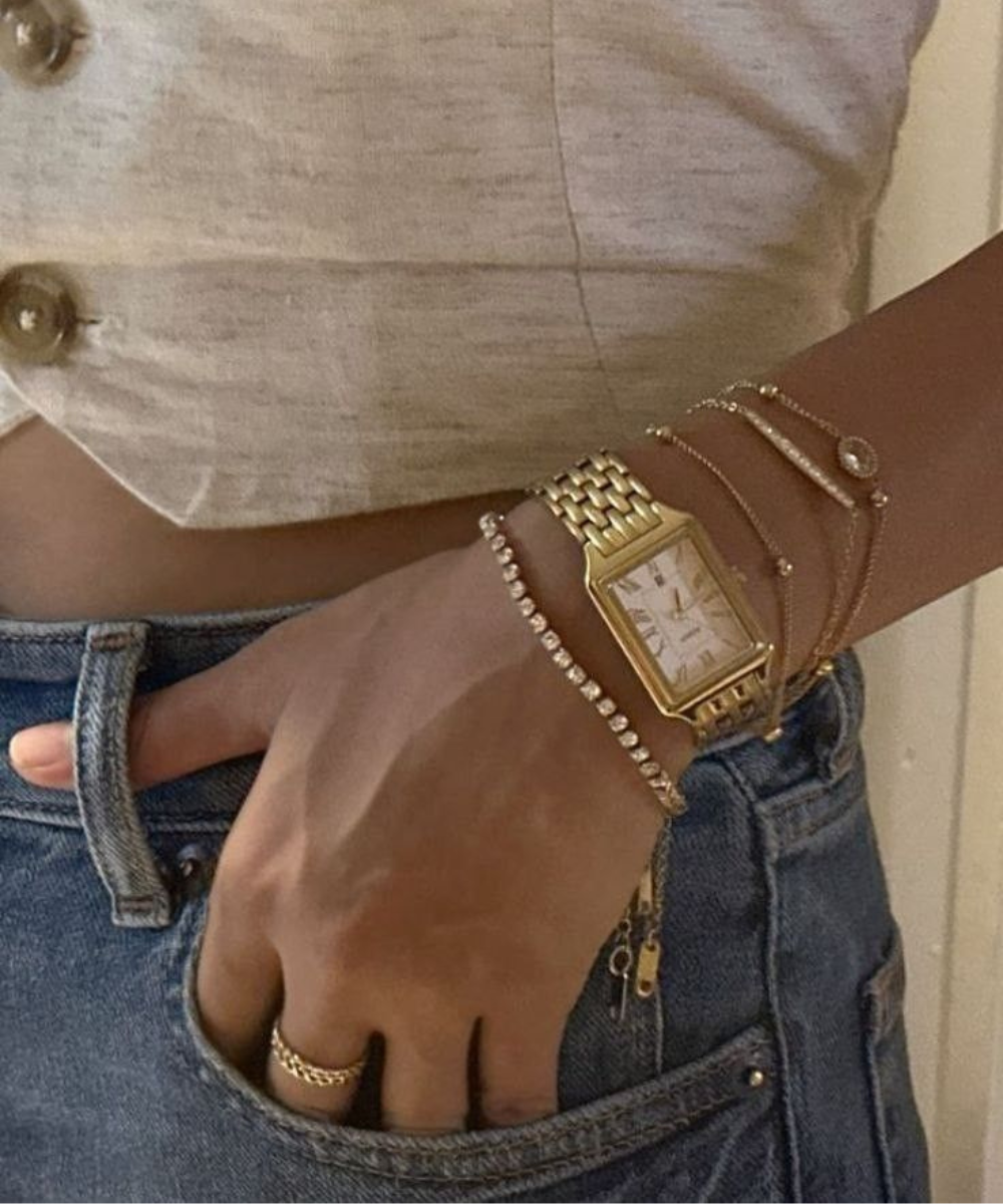 Relógio feminino dourado com pulseira de elos retangulares, combinado com pulseiras delicadas e jeans azul, estilo casual