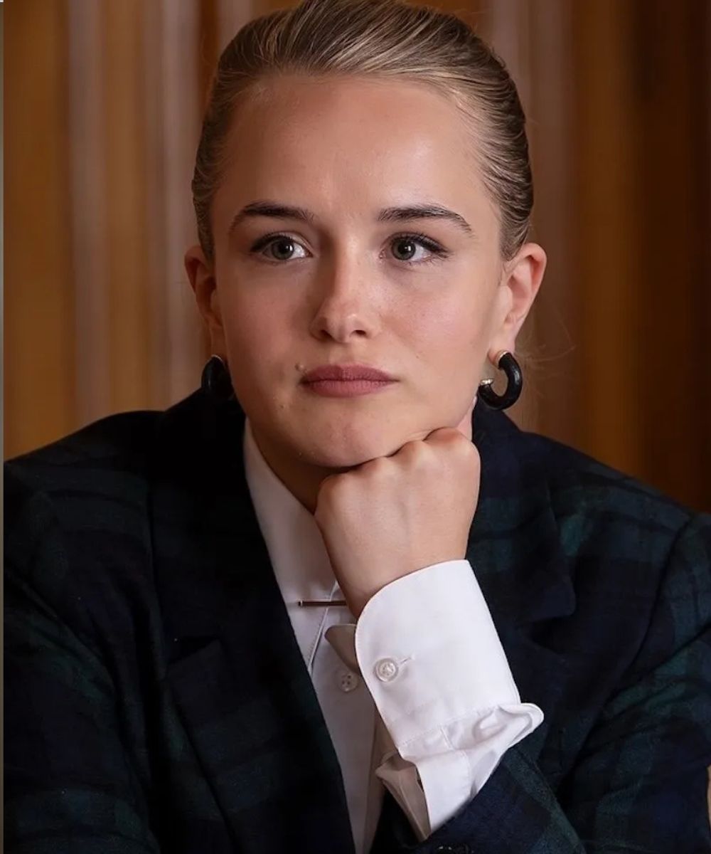 Na quarta temporada de Industry, modelo posa pensativa com blazer escuro xadrez e camisa branca de punhos largos, clima