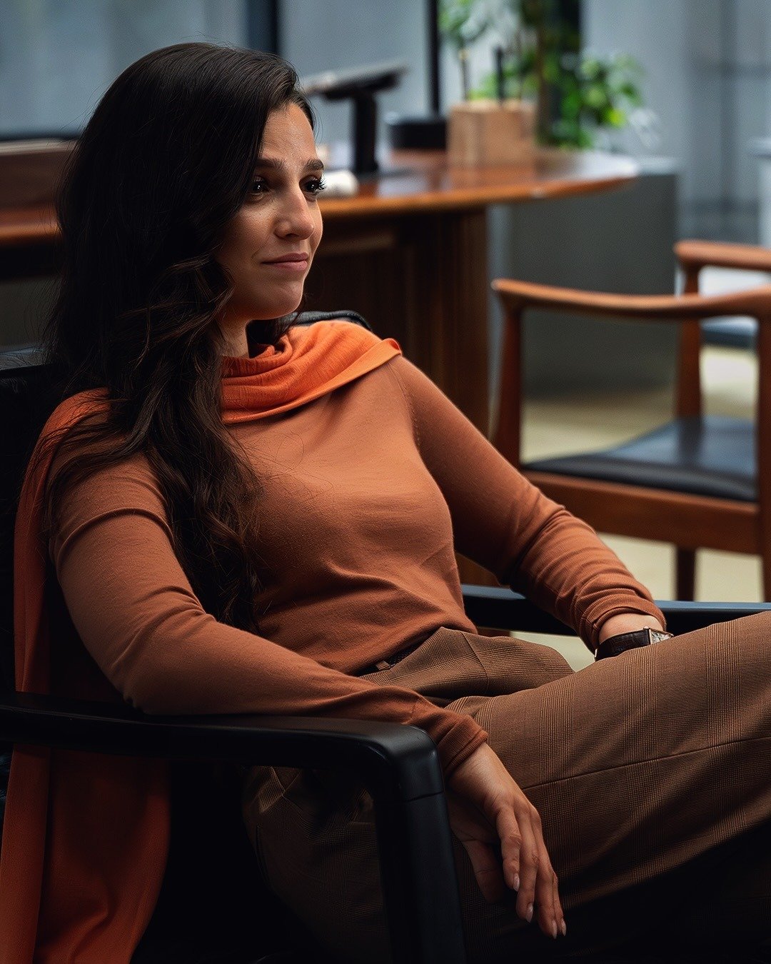 Mulher sentada com postura confiante usa blusa de malha laranja e calça xadrez marrom em ambiente corporativo sóbrio na