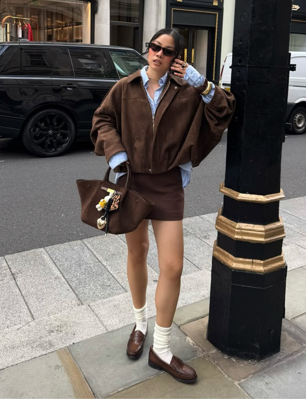 Modelo posa na rua com jaqueta bomber de camurça marrom oversized, mini saia combinando, camisa azul clara, mocassim e meias.