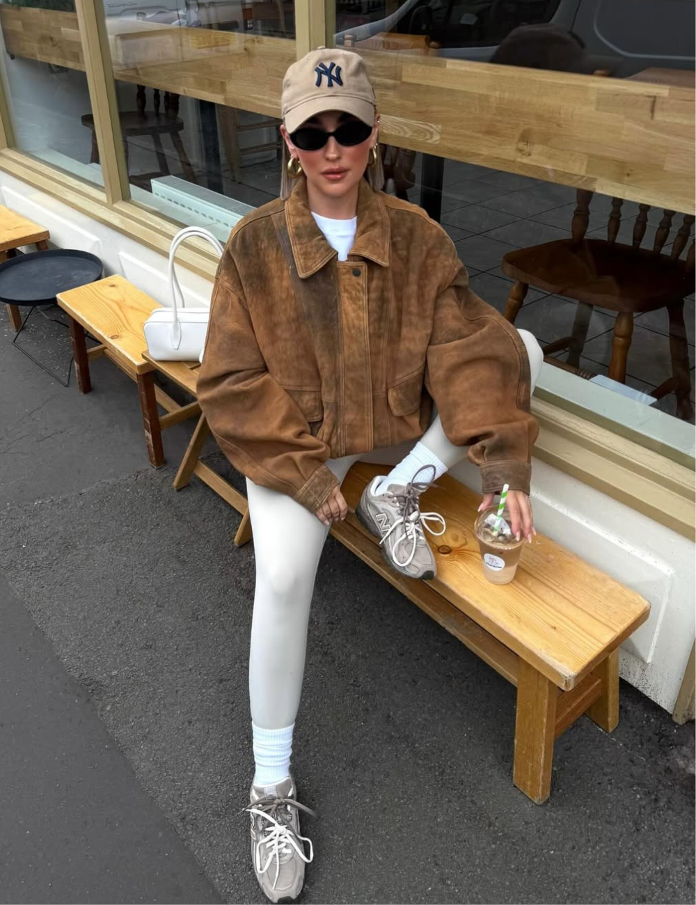 Modelo senta casualmente em banco de madeira usando jaqueta bomber de camurça marrom oversized, legging branca e boné bege.