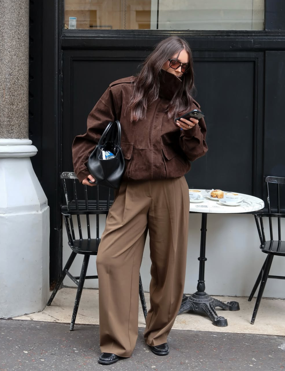 Modelo posa olhando o celular com jaqueta bomber de camurça marrom escuro oversized, calça ampla bege e bolsa preta.