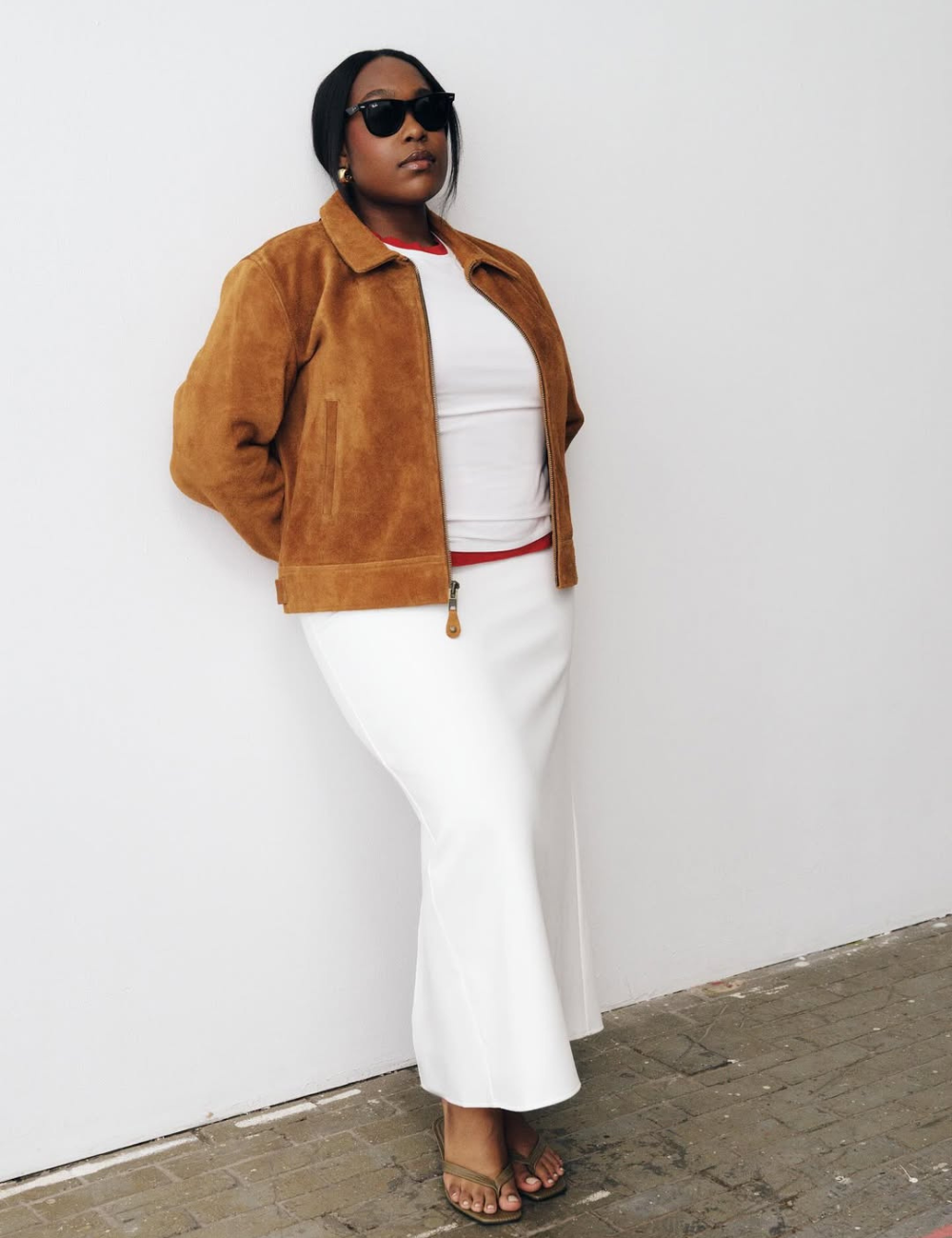Modelo posa com jaqueta bomber de camurça caramelo, combinada a saia longa branca e camiseta casual.