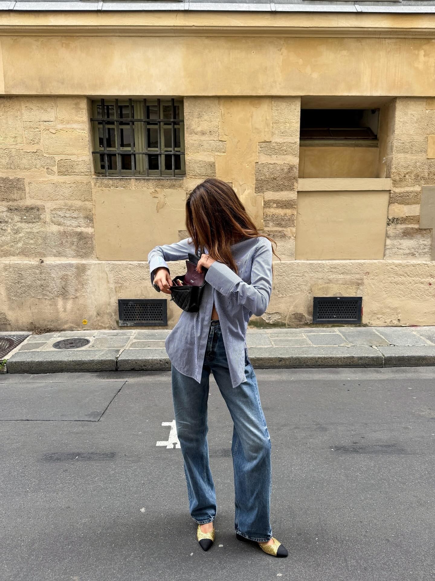 Modelo ajusta bolsa preta enquanto veste jeans azul de modelagem reta e sapatilha bicolor dourada com bico preto