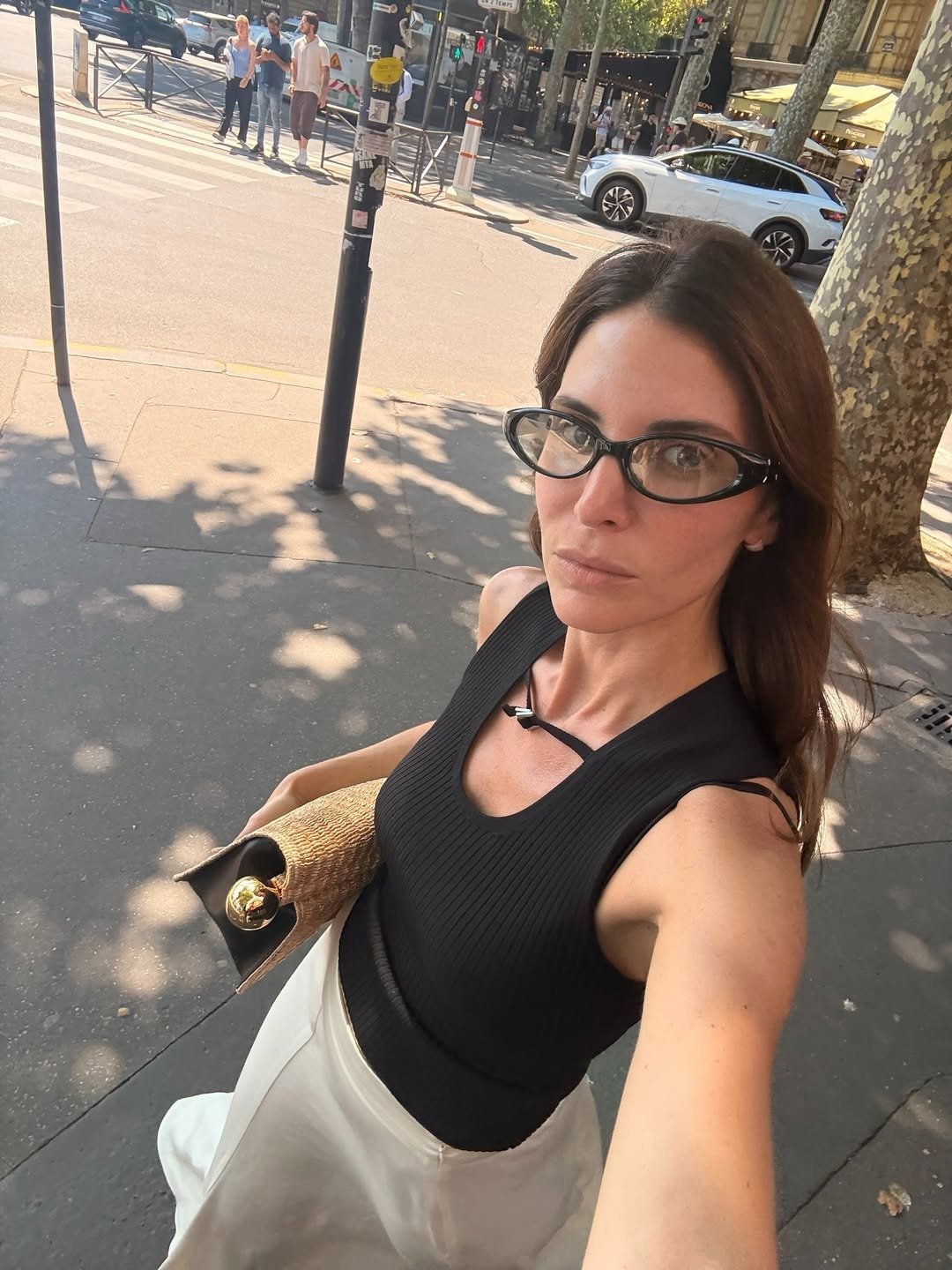 Modelo posa na rua com regata preta canelada de modelagem ajustada, combinada a saia branca fluida e bolsa de palha, em