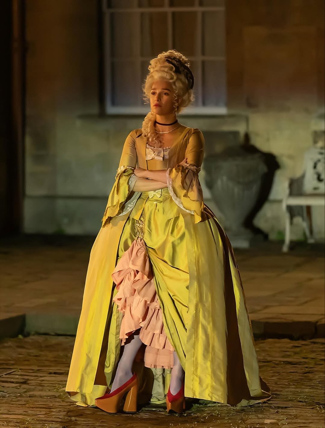 Personagem da quarta temporada de Industry veste vestido amarelo com saia volumosa e babados rosa, posa com braços cruzados