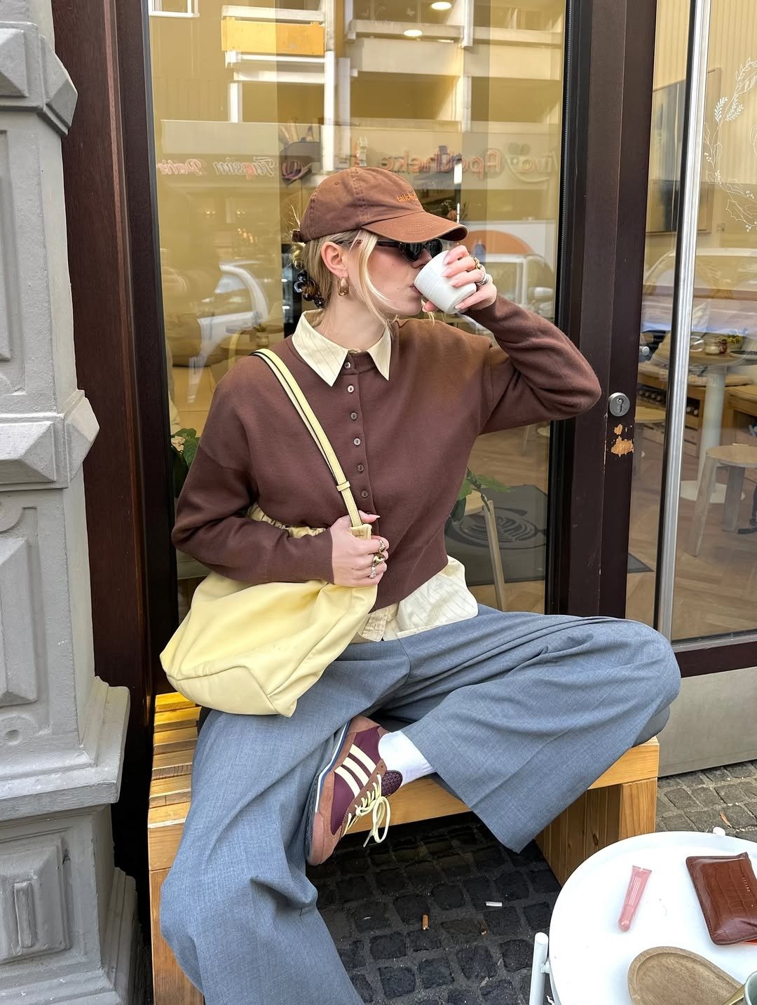 Modelo senta casualmente em banco de madeira, veste blusa marrom, camisa bege, calça cinza ampla e tênis retrô vinho com