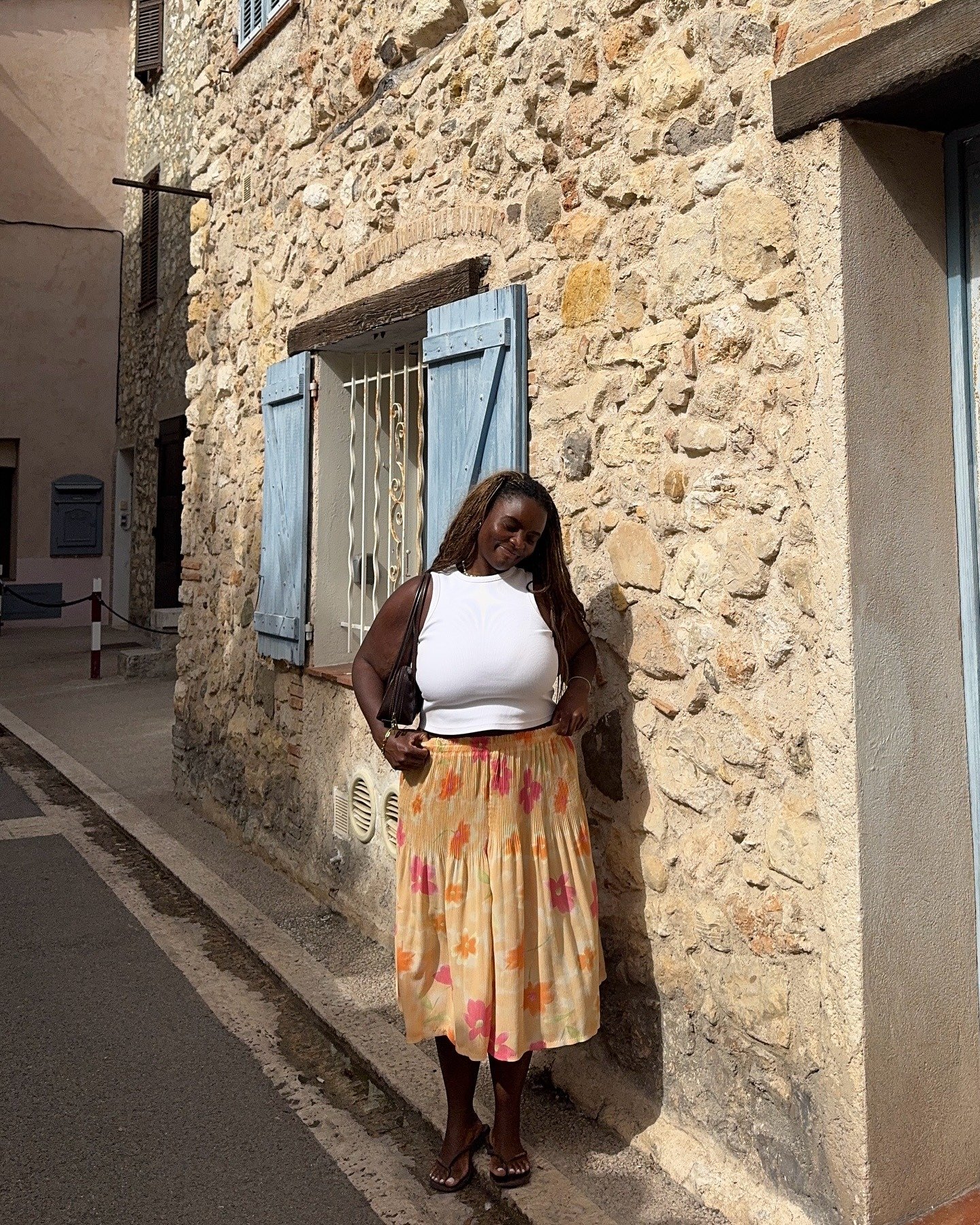 Modelo posa sorrindo e segura a saia midi plissada com estampa floral em tons de laranja e rosa, combinada com regata branca