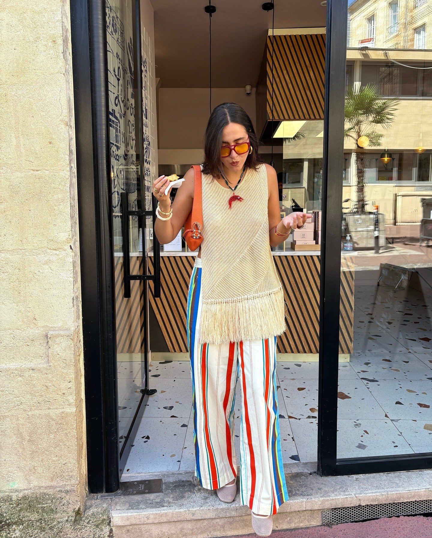 Mulher posa com blusa bege de tricô com franjas, calça ampla listrada em laranja, azul e branco e bolsa laranja vibrante