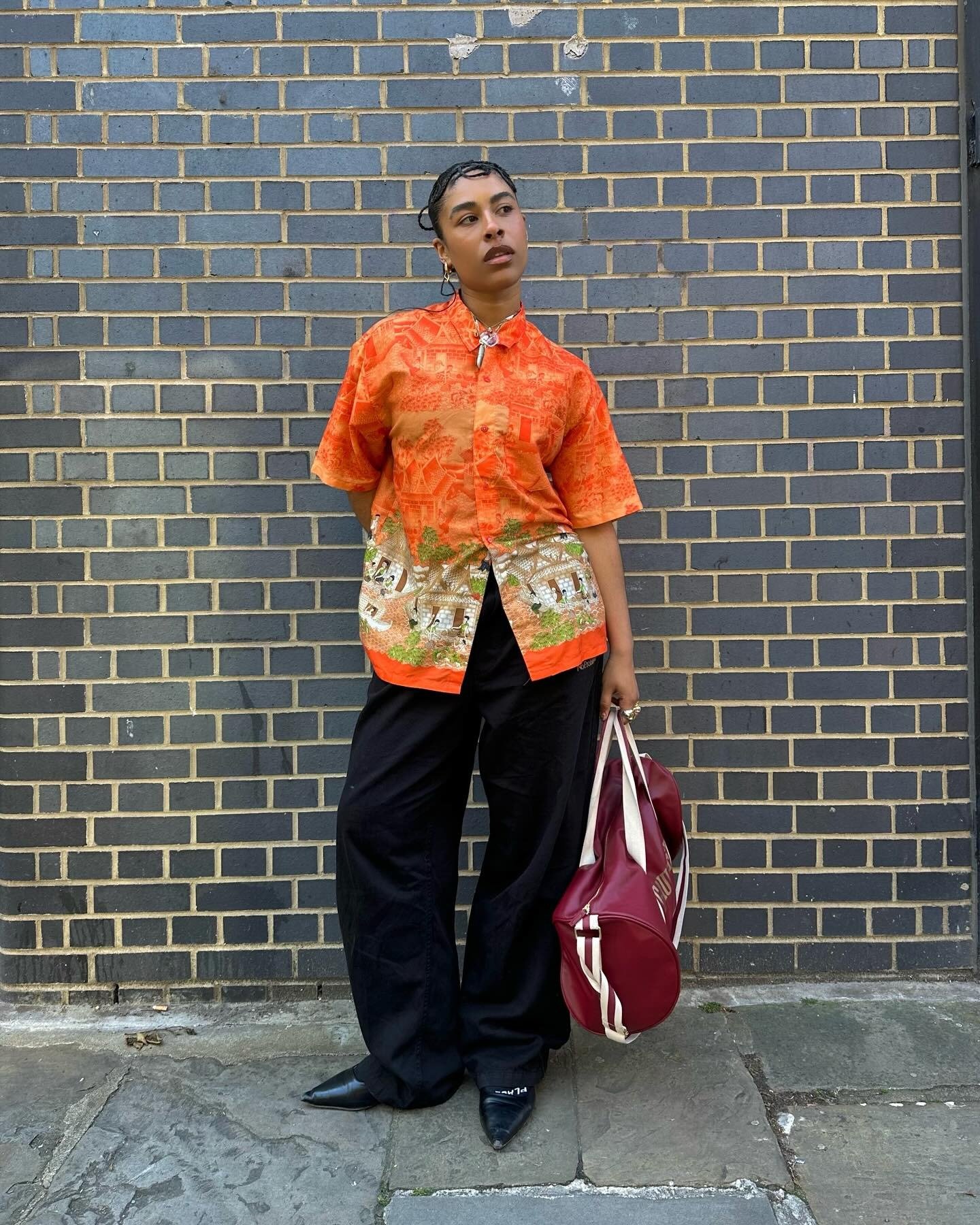 Modelo posa com camisa laranja estampada em tecido leve, calça preta ampla e bolsa vinho, criando looks com laranja modernos