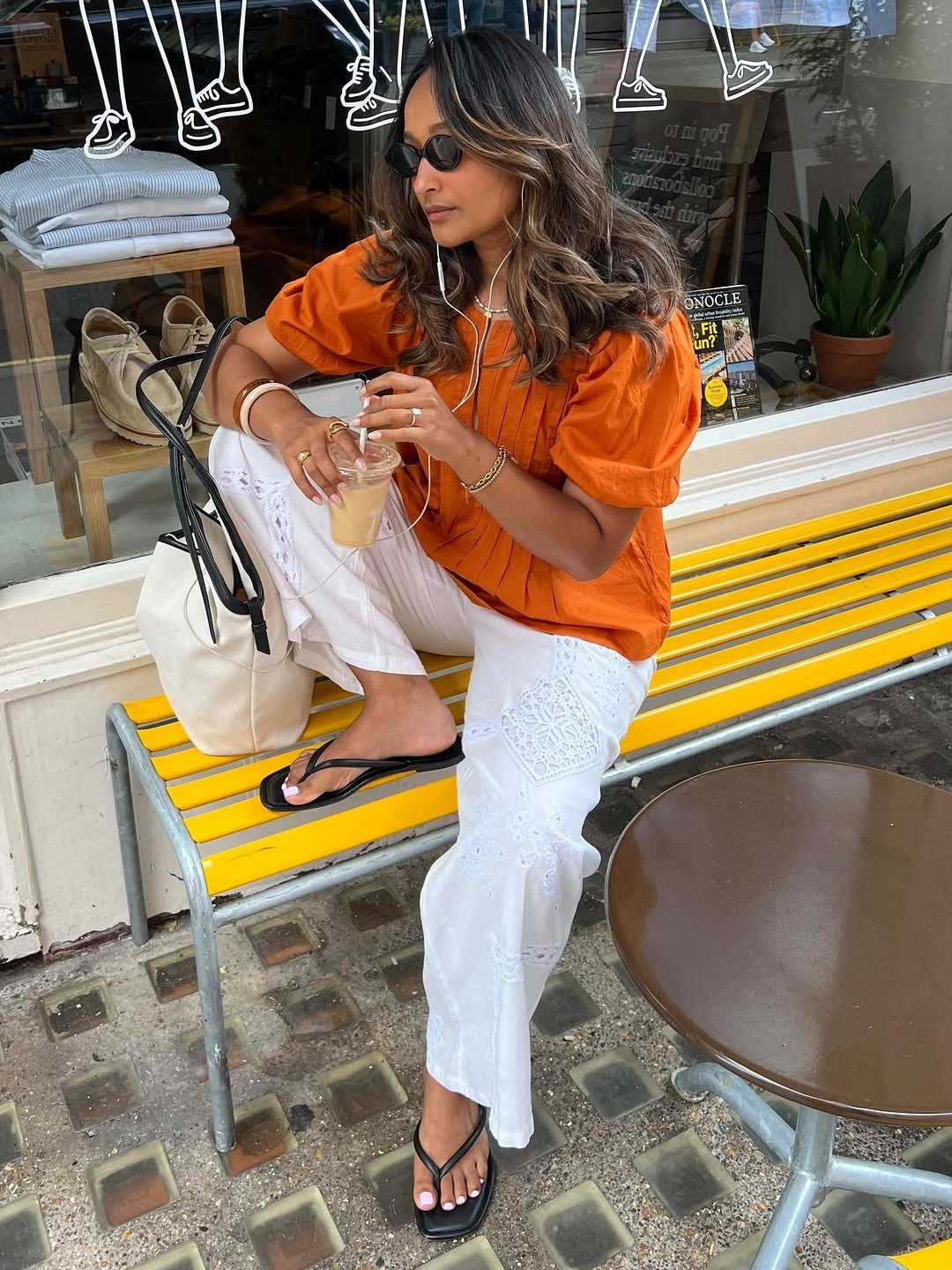 Mulher senta em banco amarelo com blusa laranja de mangas bufantes e calça branca bordada, em look com laranja casual