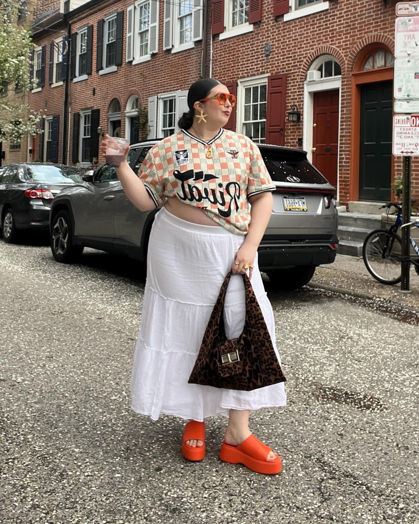 Modelo posa na rua com blusa cropped laranja xadrez, saia branca fluida, sandálias plataforma laranja e bolsa animal print