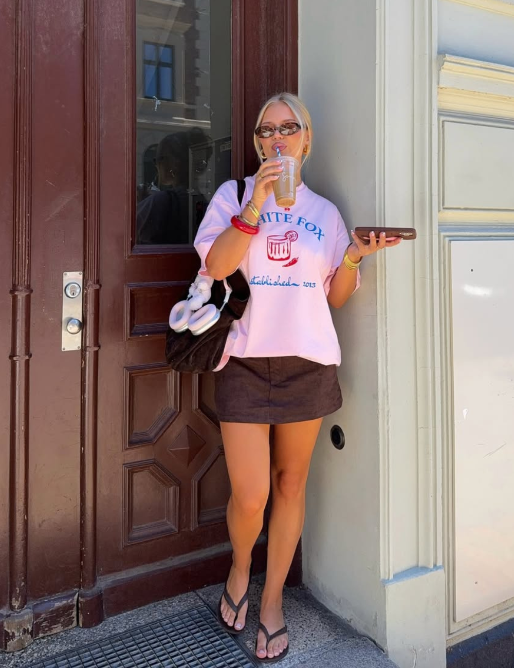 Modelo posa encostada em parede com look casual de minissaia marrom em tecido texturizado e camiseta rosa over.