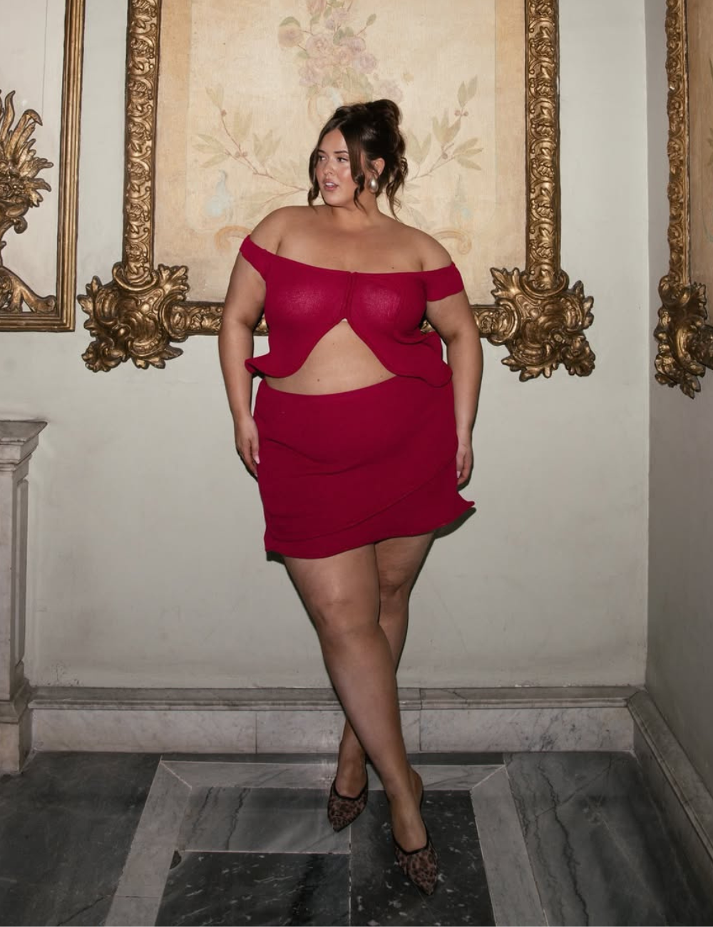 Modelo posa com look com minissaia vermelha de tecido estruturado e top ombro a ombro, em ambiente clássico e elegante.