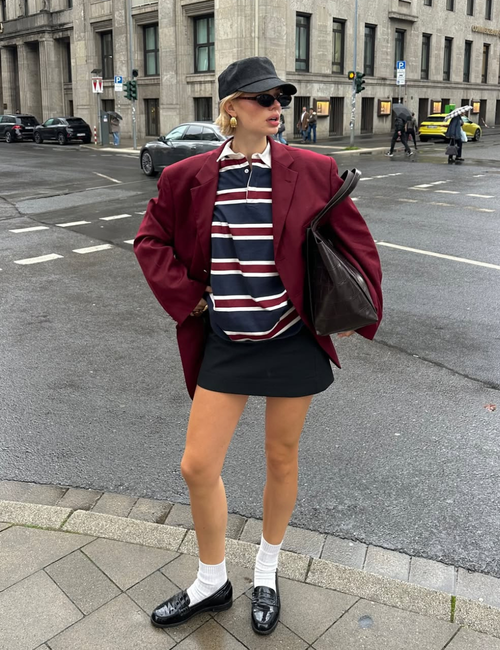 Modelo posa na rua com look urbano que combina minissaia preta reta, blazer vinho oversized e camisa polo listrada.