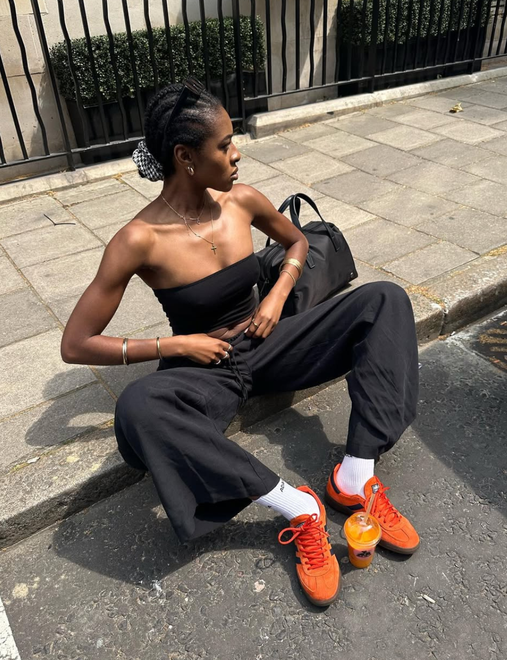 Modelo senta no meio-fio com postura relaxada, veste top faixa e calça pantacourt preta, tênis laranja vibrante e meias.