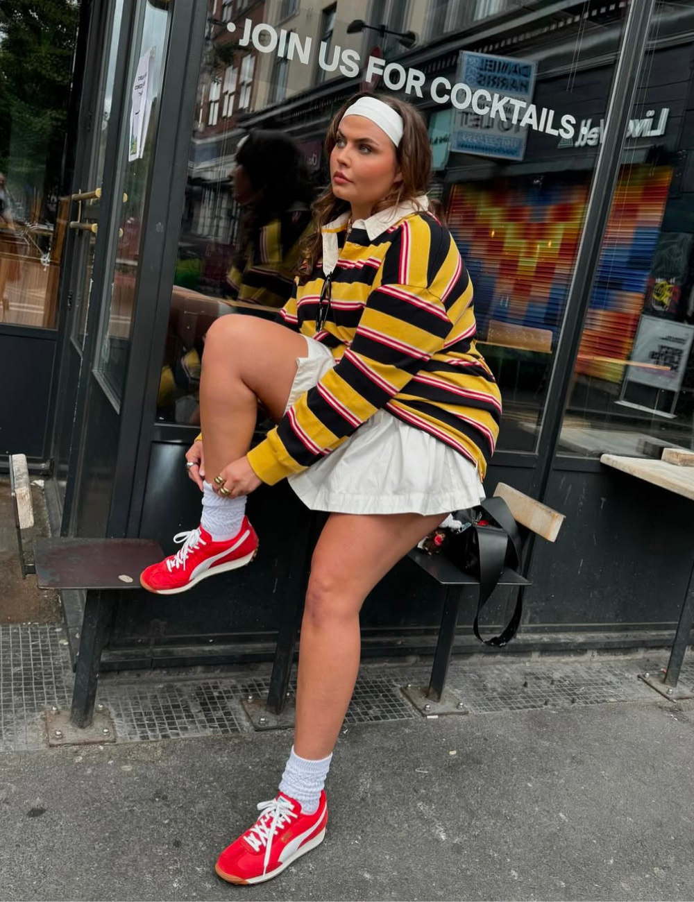 Modelo senta ajustando tênis colorido vermelho com branco, combinando com blusa listrada amarela, preta e vermelha e saia.