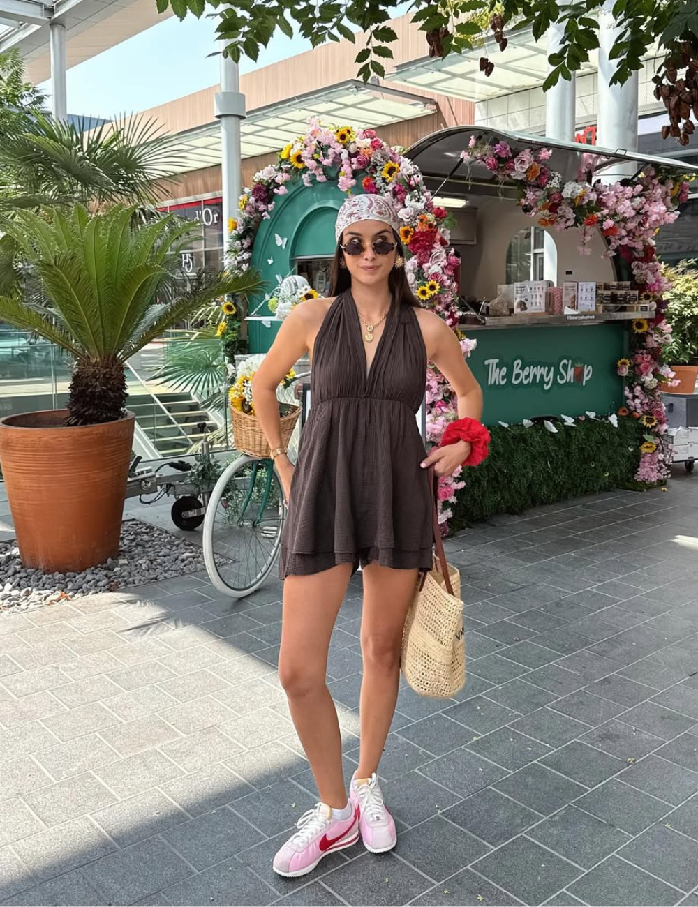 Modelo posa com vestido curto preto de tecido leve, tênis rosa com detalhes vermelhos, lenço estampado e acessórios.