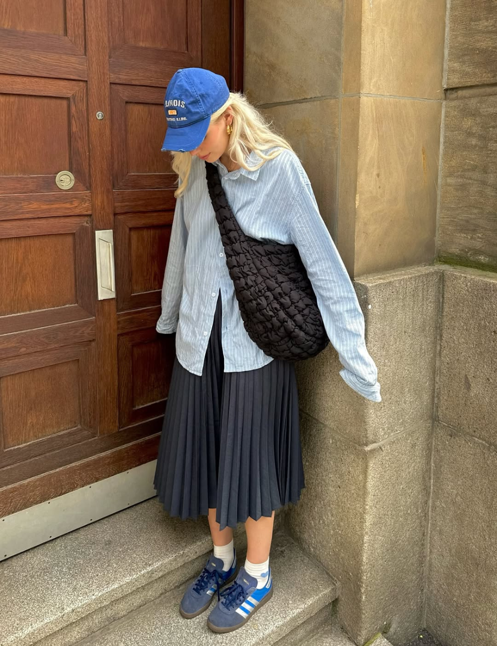 Modelo posa com camisa listrada azul clara oversized, saia plissada preta midi e tênis colorido azul com branco.