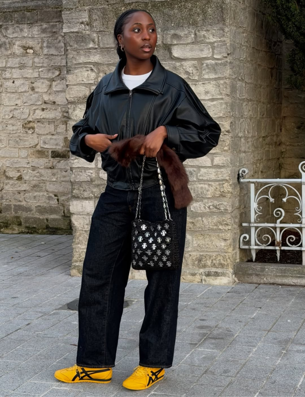 Modelo posa com jaqueta preta de couro sintético, jeans escuro de modelagem reta e tênis amarelo vibrante.
