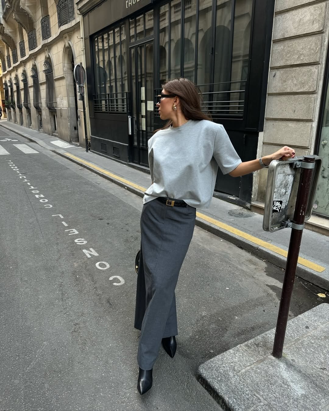 Modelo posa com looks lindos em saia longa cinza, blusa oversized clara, cinto preto e bota de couro, em clima urbano