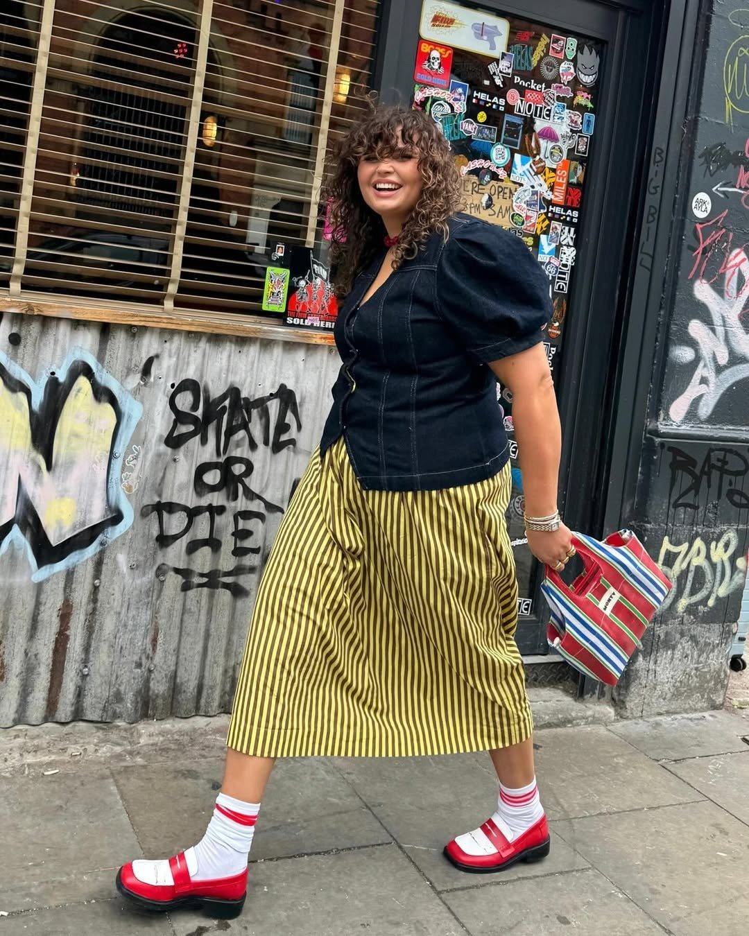 Modelo caminha sorrindo com looks lindos em jaqueta jeans preta de mangas bufantes, saia midi listrada amarela e preta