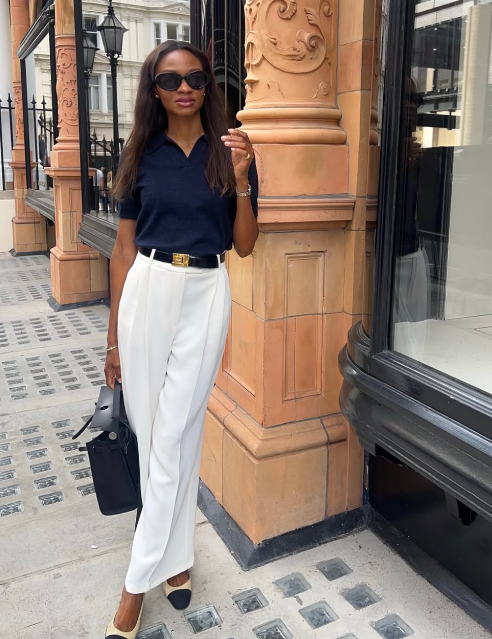 Modelo posa com looks novos e bonitos em calça branca de alfaiataria, blusa azul marinho, cinto preto e bolsa estruturada.