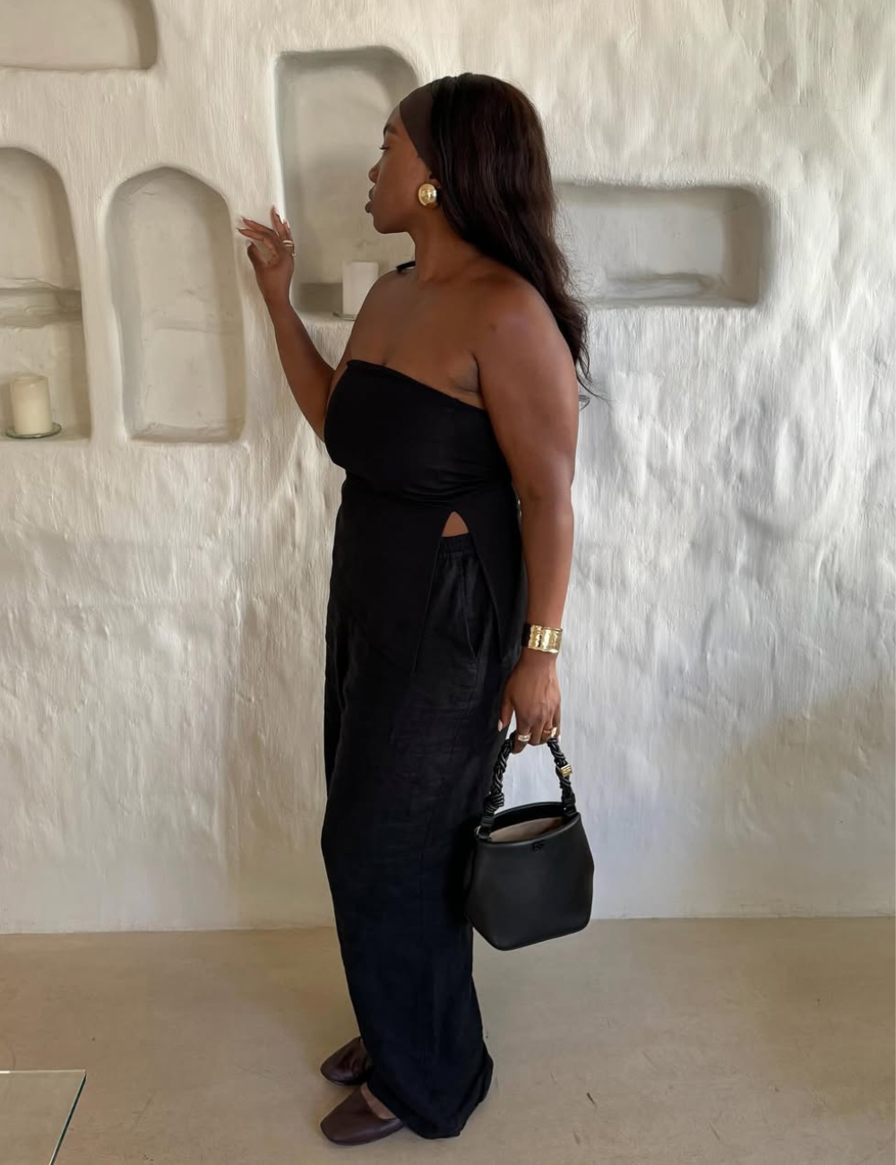 Mulher posa de perfil com vestido longo preto tomara que caia em linho, bolsa preta e acessórios dourados.