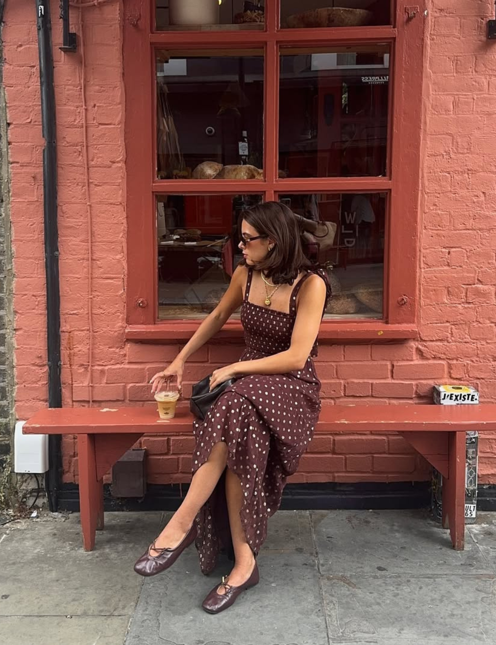 Mulher sentada em banco vermelho usa vestido marrom de alças com estampa de bolinhas claras e sapatilhas combinando.