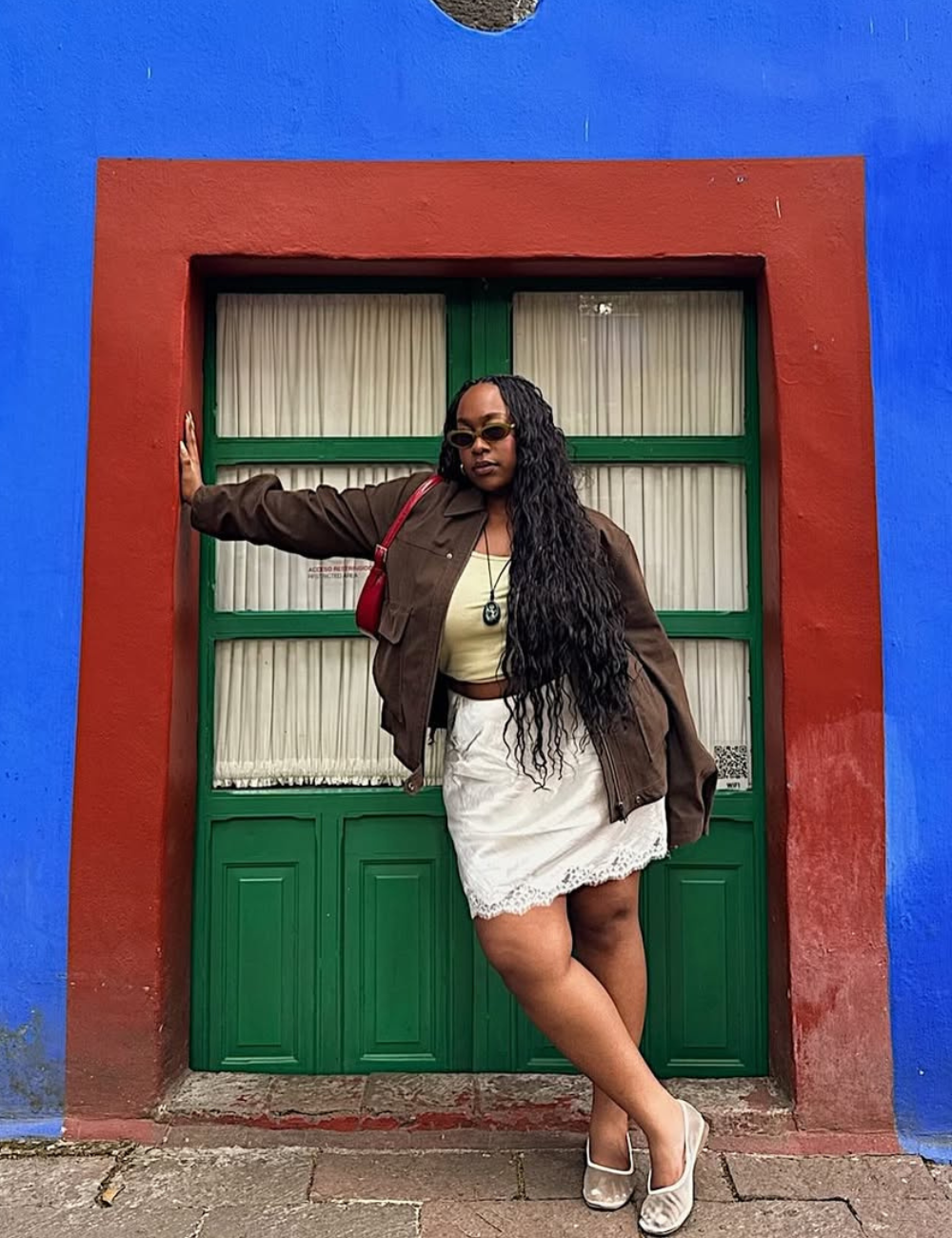 Modelo posa com looks novos e bonitos usando jaqueta oversized marrom, top amarelo, saia branca rendada e bolsa vermelha.