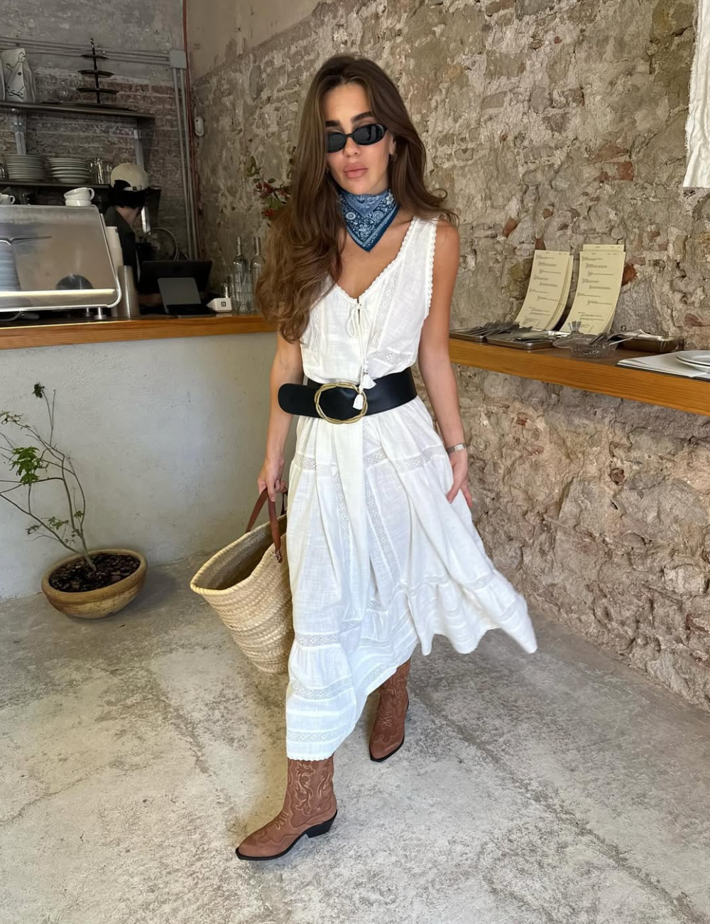 Modelo posa com vestido branco fluido de algodão, cinto largo preto, botas caramelo, lenço azul e bolsa de palha.