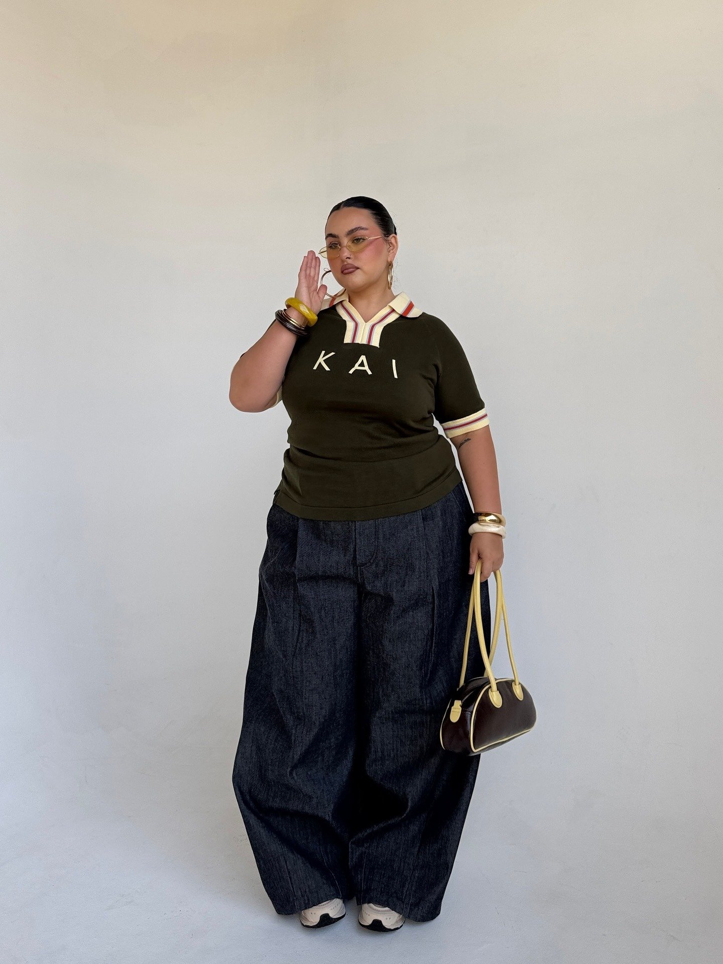 Modelo posa com postura confiante usando polo verde-musgo com gola listrada, calça jeans ampla e acessórios