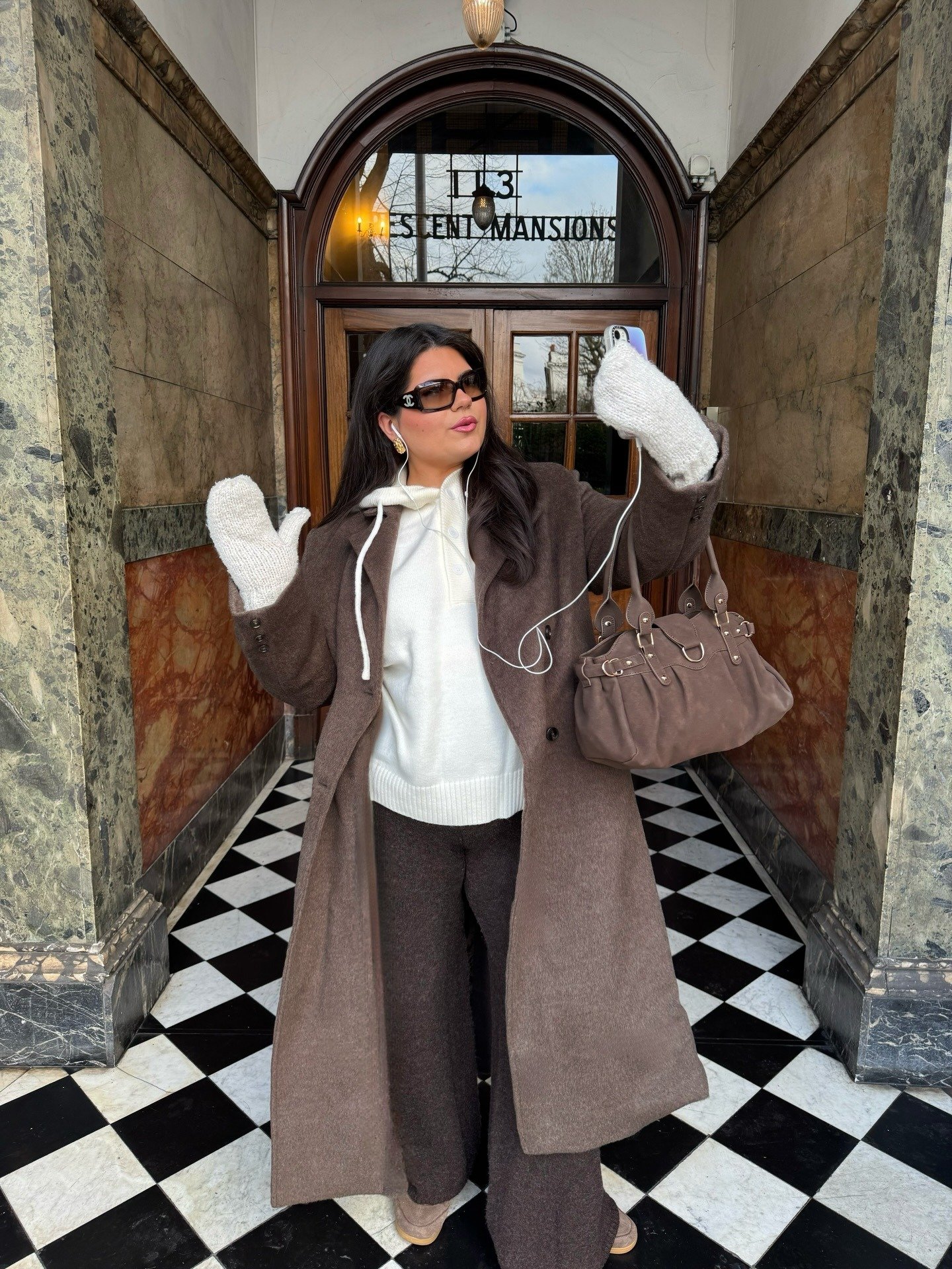 Mulher posa com luvas de tricô bege, casaco longo marrom, calça ampla e suéter branco em ambiente clássico e elegante.