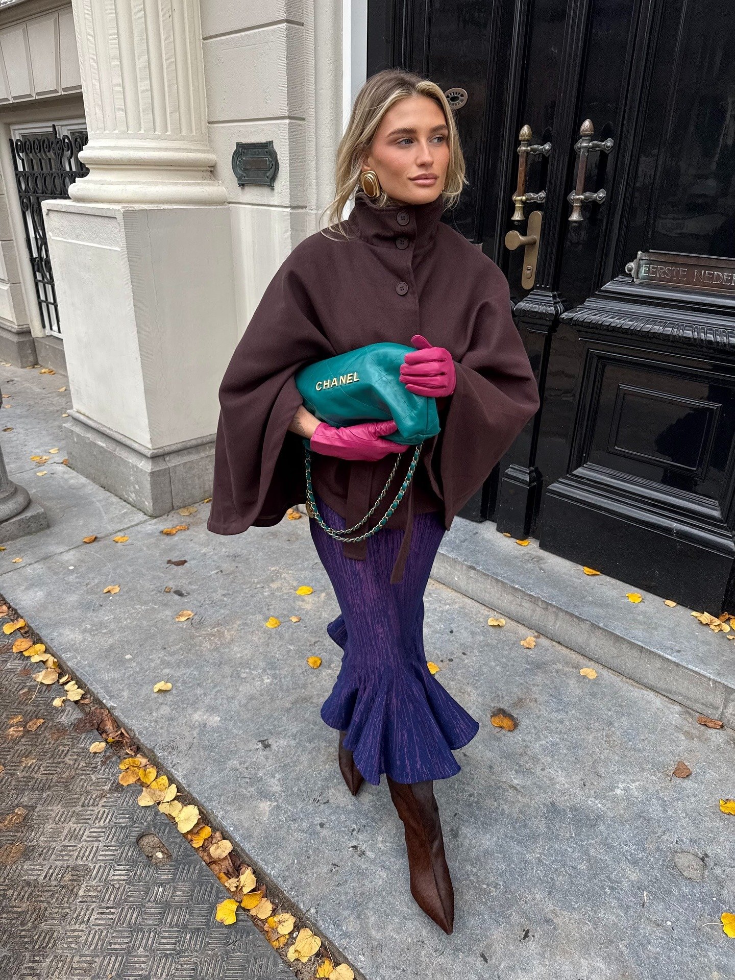 Modelo posa com luvas de couro rosa vibrante, capa marrom oversized, saia azul texturizada e bota marrom pontuda, segurando