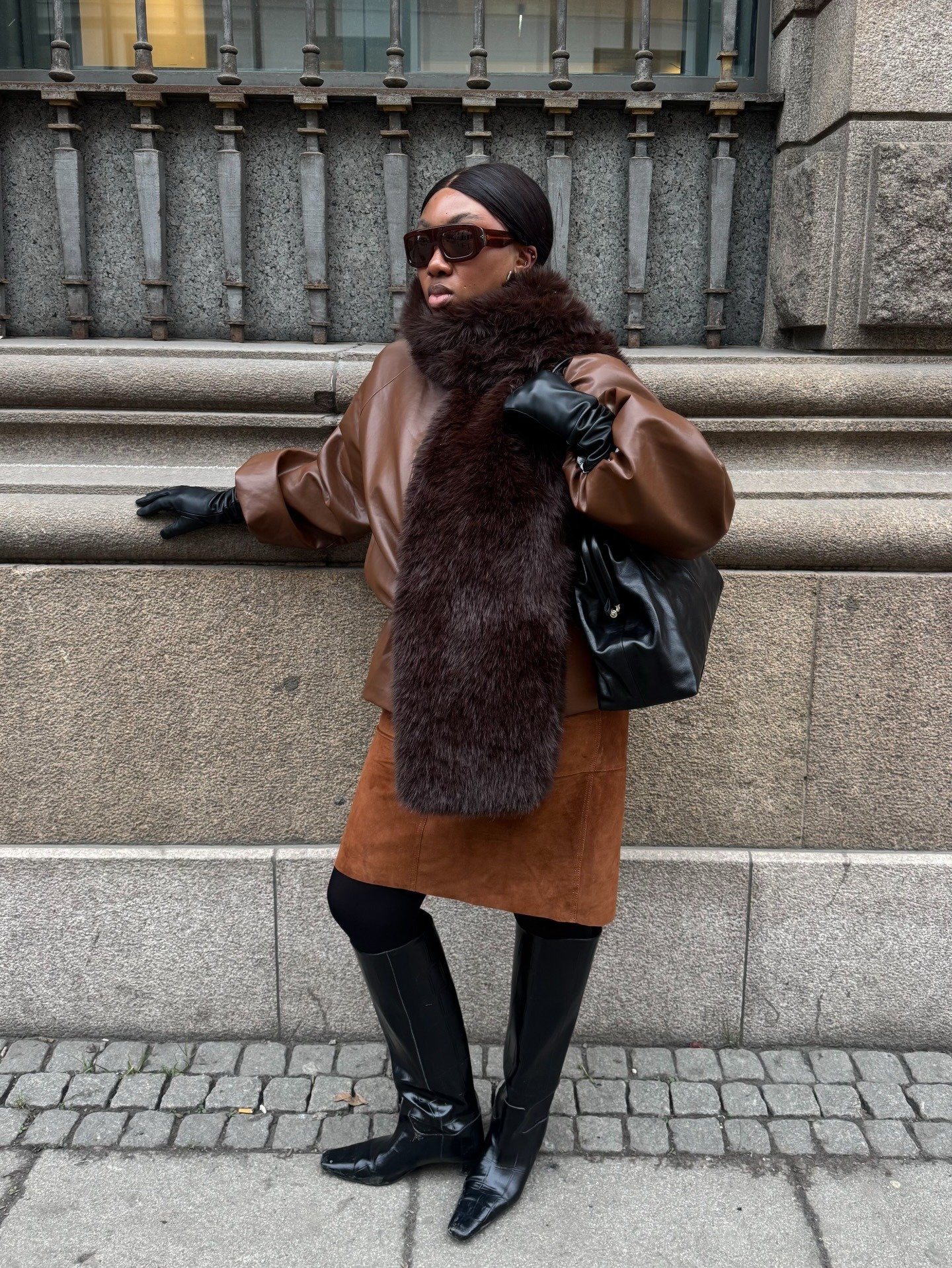 Modelo posa com luvas pretas de couro, casaco marrom oversized, saia camurça caramelo, botas longas pretas e cachecol de