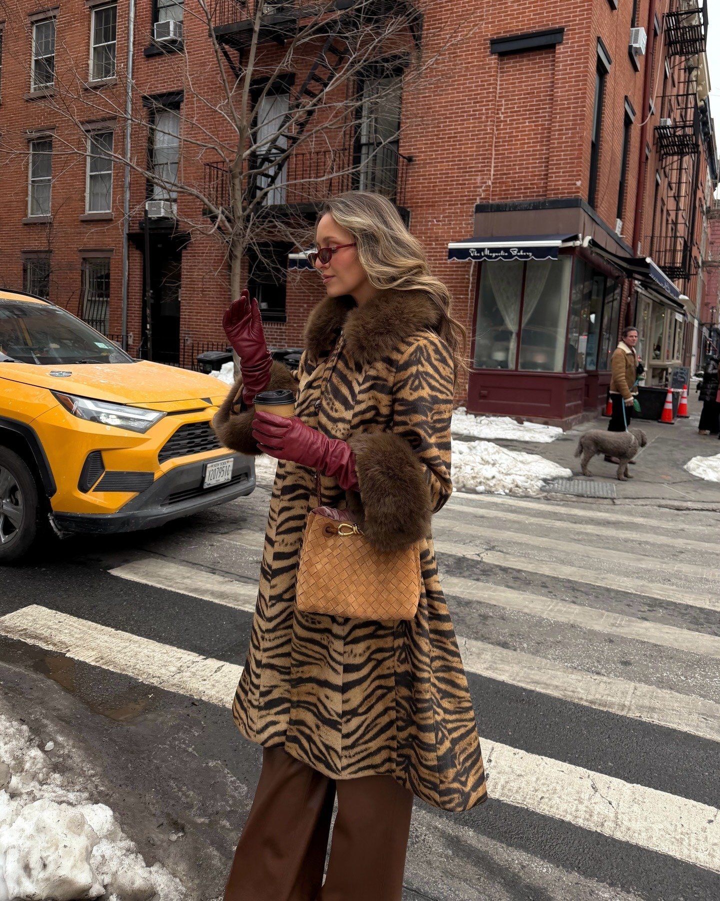 Mulher posa na rua com luvas de couro vinho, casaco animal print com peles marrons e bolsa caramelo trançada, clima urbano