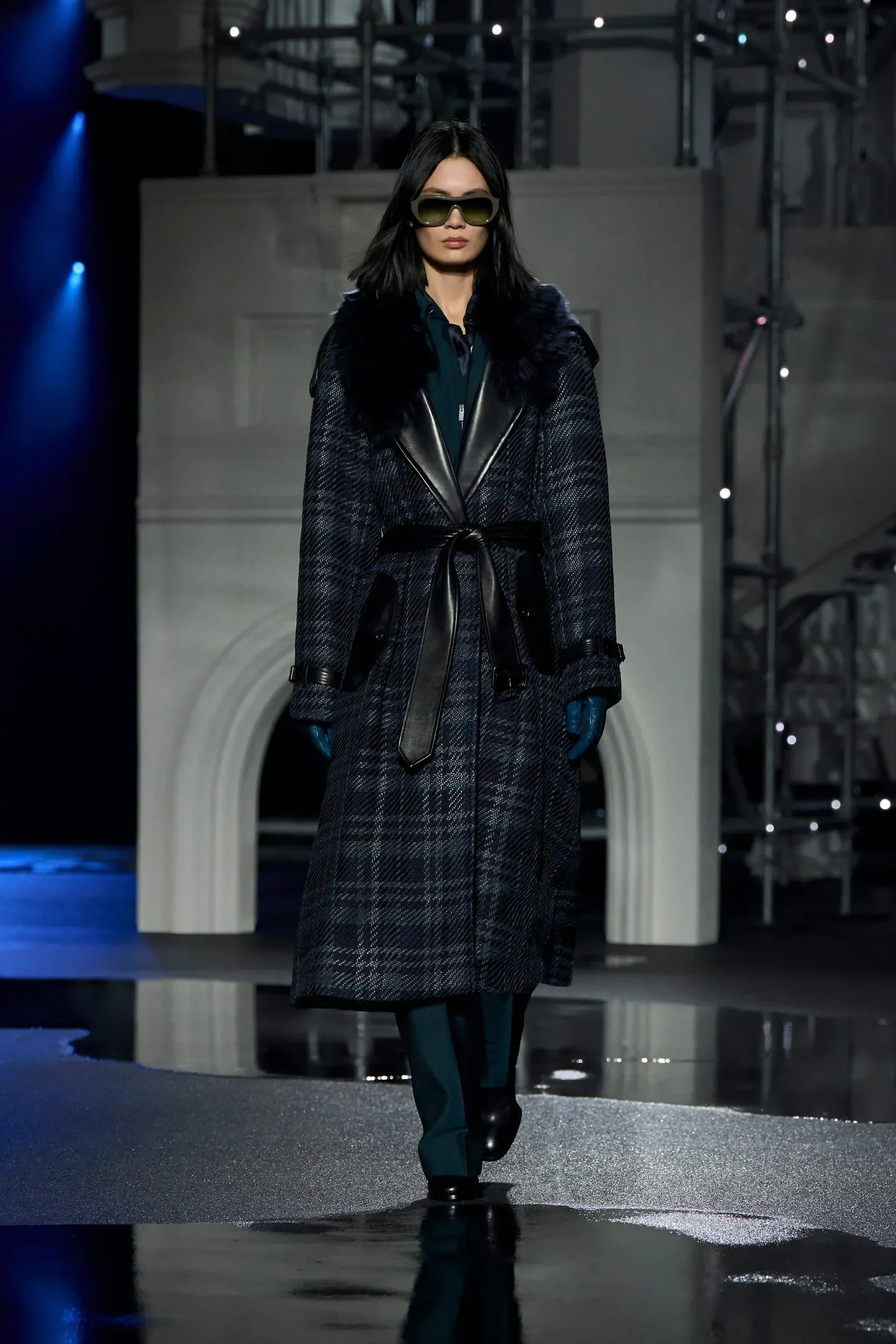 Modelo caminha com postura firme usando luvas de couro azul, sobretudo xadrez escuro com detalhes em preto e gola de pelo