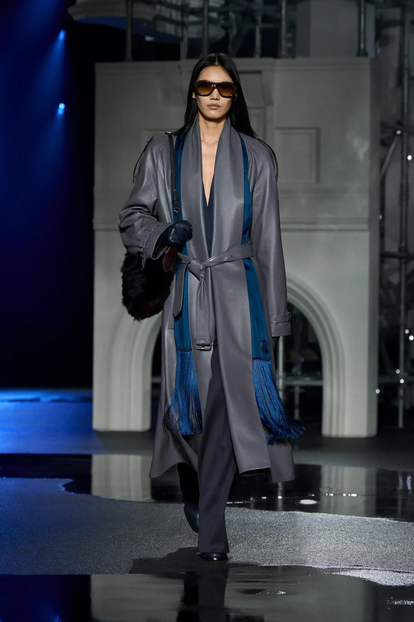 Modelo caminha com postura confiante usando luvas de couro pretas que complementam sobretudo cinza com detalhes em azul  
