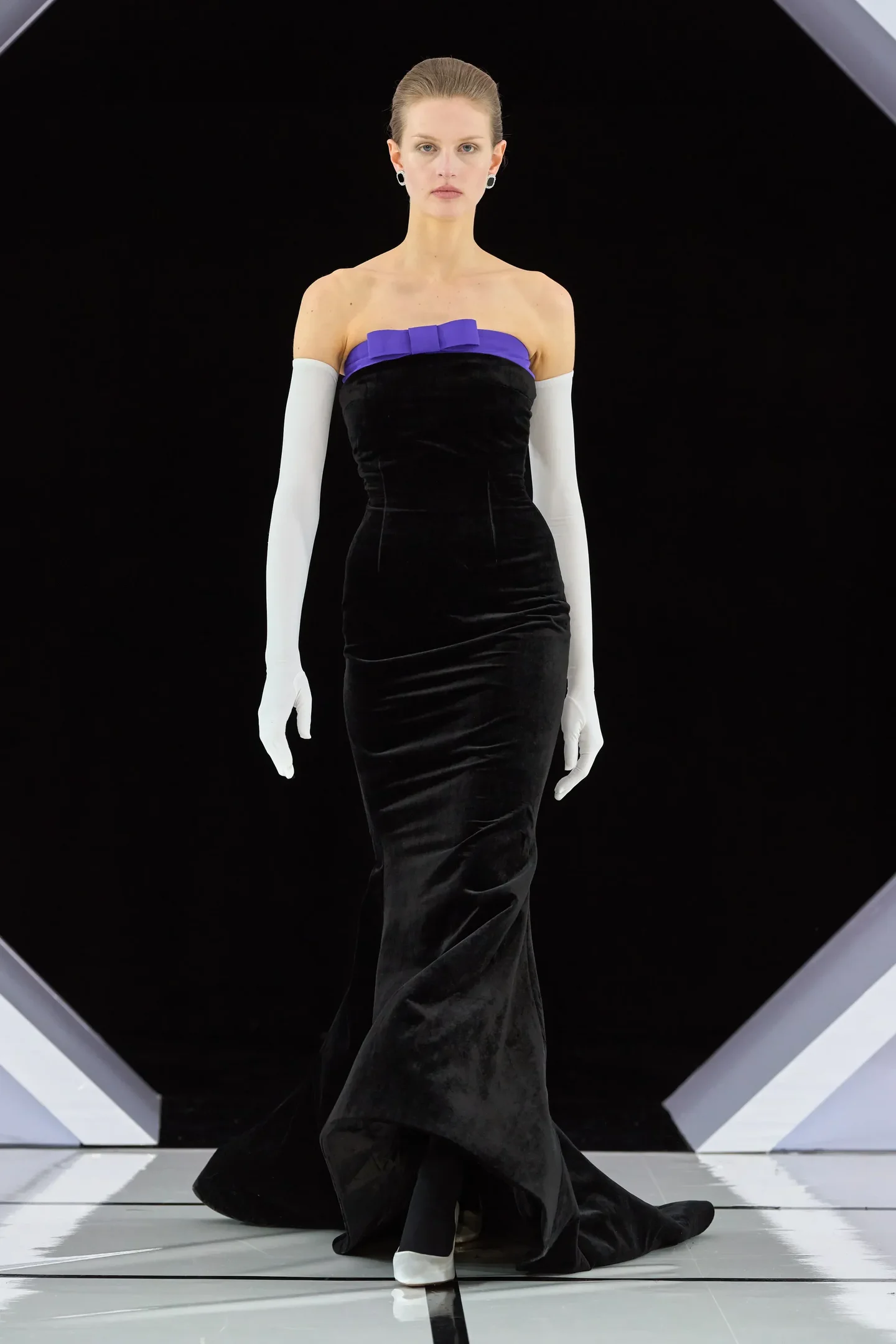 Modelo caminha com postura firme usando vestido longo justo de veludo preto com detalhe roxo no busto e luvas longas brancas