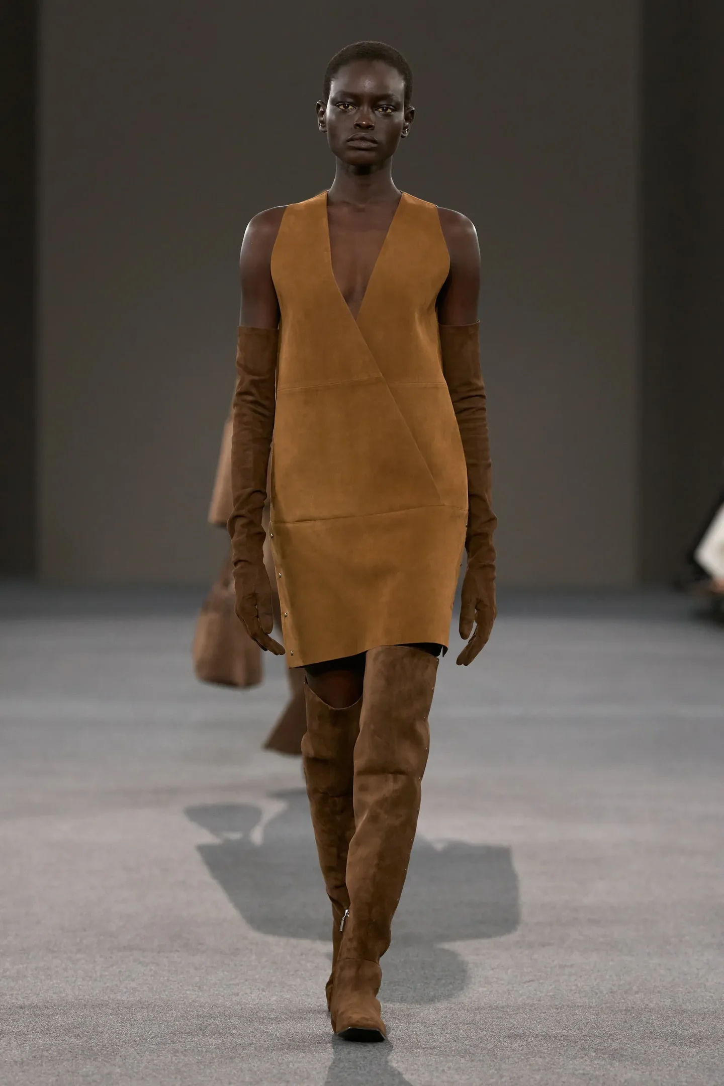 Modelo caminha com postura firme usando vestido marrom em suede, luvas longas e botas over the knee do mesmo material 