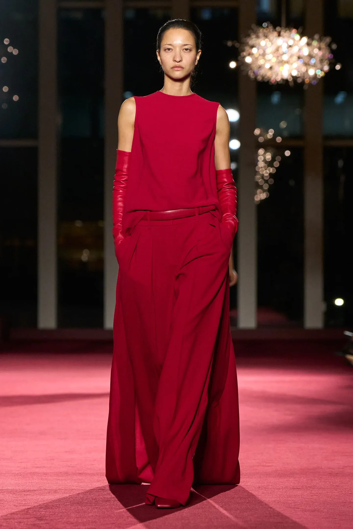 Modelo caminha com postura firme e expressão séria usando conjunto vermelho fluido com calça ampla, top sem mangas e luvas