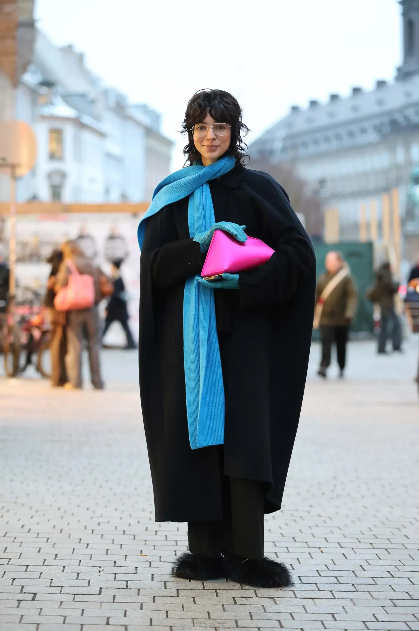 Mulher posa com luvas de couro azul, cachecol longo e bolsa pink, combinando com casaco preto oversized em rua urbana.