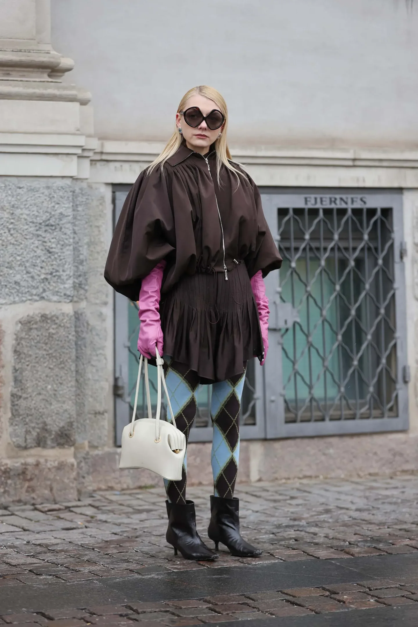 Modelo posa com luvas longas rosa vibrante, vestido marrom de mangas bufantes, meia-calça xadrez azul e preta e bota preta,