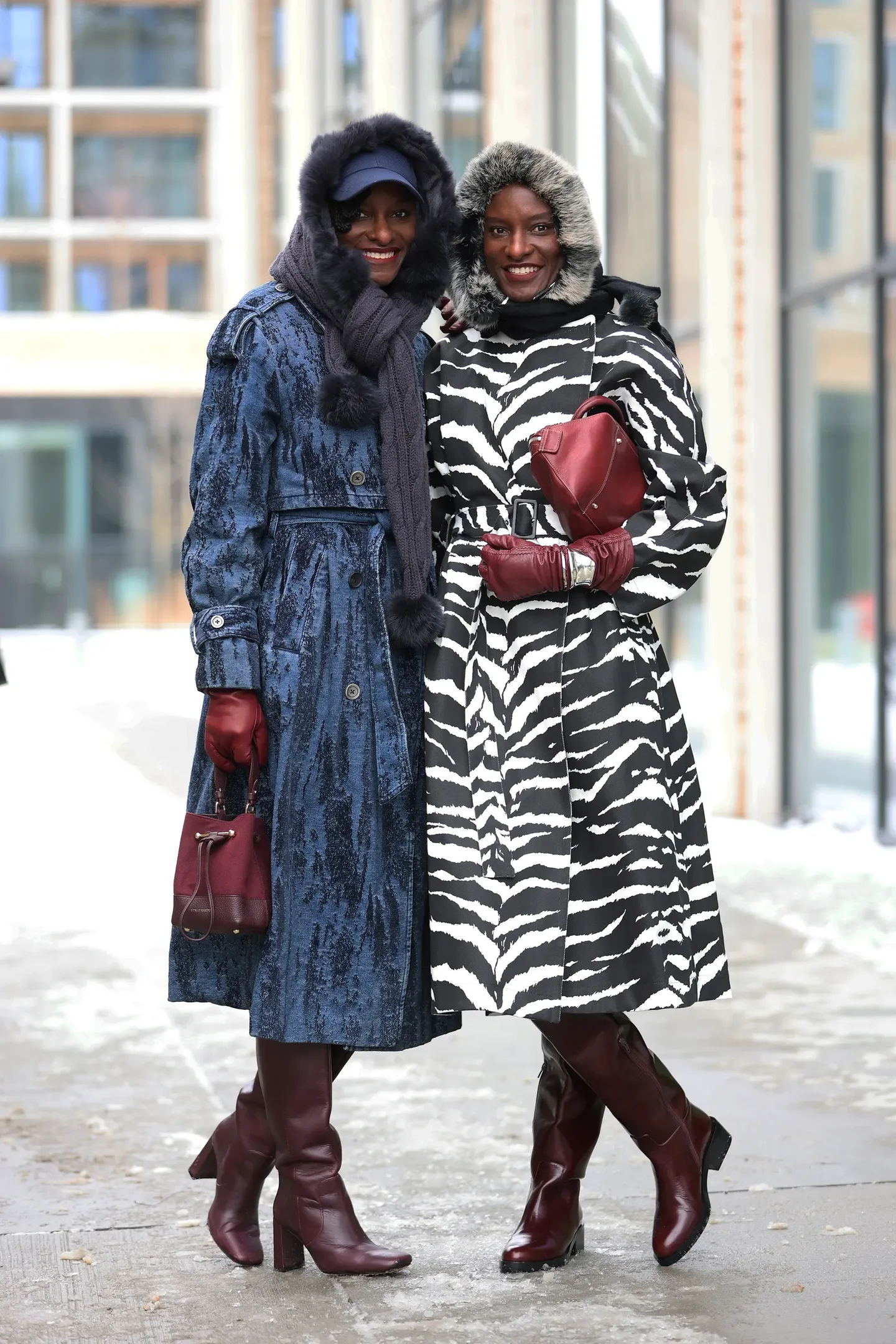 Duas mulheres posam sorrindo com luvas de couro vinho, casacos longos em azul texturizado e estampa zebra, botas 